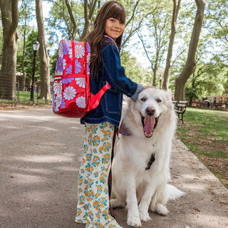 State Bags Kane Double Pocket Backpack - Ruby Floral Girl with a colorful backpack and floral pants standing next to a white dog in a park.