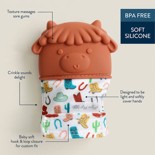 Itzy Ritzy Teething Mitt - Highland Cow Silicone teething toy with cowboy design and text highlighting features on a beige background