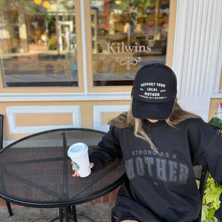 Support Your Local Mother® Trucker Hat - Black
Person sitting at a table outside a business named Kilwins, wearing a cap and sweatshirt with text.