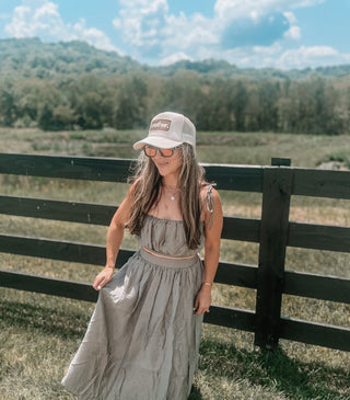 Mother Patch Trucker Hat - Sand
Woman in a gray dress and cap standing in front of a wooden fence with a scenic background