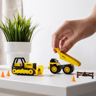 Schylling Tonka Mini Metals 2 Pack 
Hand interacting with Tonka toy construction vehicles on a white surface with a plant in the background.