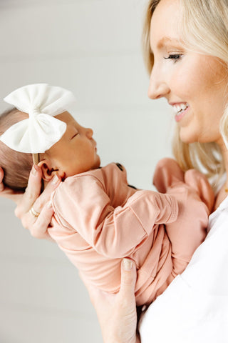 Lou Lou and Company Top and Bottoms - Abby
Woman holding a baby wrapped in pink fabric with a white bow on a plain background