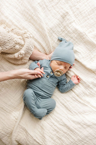 Lou Lou and Company Ribbed Top and Bottoms - Ford
Newborn baby in a blue outfit and hat lying on a soft beige blanket.