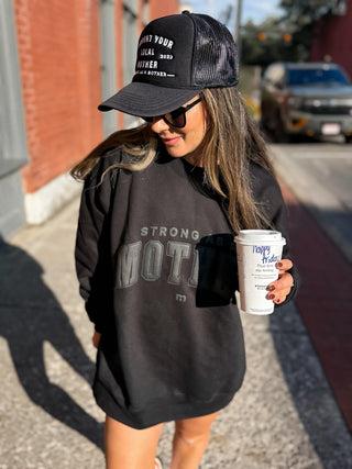 Support Your Local Mother® Trucker Hat - Black
Woman wearing a black cap and hoodie, holding a coffee cup on a street.