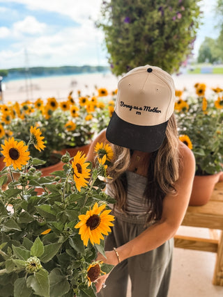 Strong As A Mother® Script Canvas Hat - Natural / Black
Woman wearing a cap with text, standing among sunflowers by a waterfront.