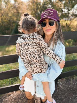 Support Your Local Mother® Trucker Hat - Burgundy
Woman holding a child outdoors with a wooden fence and trees in the background