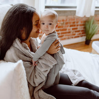 Bonsie Footie - Fog
Woman holding a baby in a cozy indoor setting with a brick wall and plant in the background.