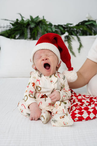 Lou Lou and Company Santa Beanie
Newborn baby wearing a Santa hat and onesie with Christmas patterns, yawning on a white couch.