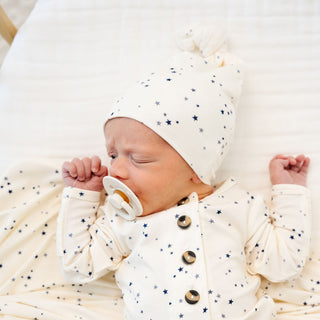 Lou Lou and Company Top Knot Hat - George
Newborn baby wearing a white outfit with star patterns and a matching hat, lying on a white blanket.