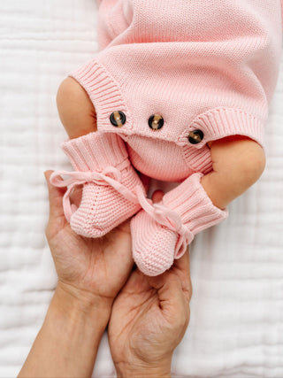 Lou Lou and Company Knit Booties - Blush
Pink knitted baby booties held by a hand with a baby in pink outfit in the background.