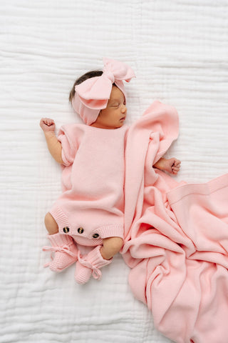 Lou Lou and Company Knit Booties - Blush
Newborn baby in pink outfit with bear design and matching headband, lying on a white textured surface.