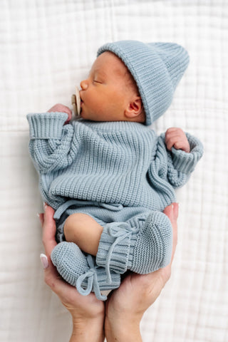 Lou Lou and Company Knit Booties - Steele
Newborn baby in matching blue knit outfit with hat and shorts, held against a white background.
