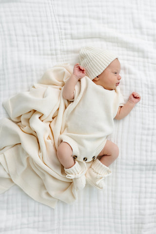 Lou Lou and Company Knit Booties - Ivory
Newborn baby wrapped in a white blanket and wearing a matching hat on a white bed.