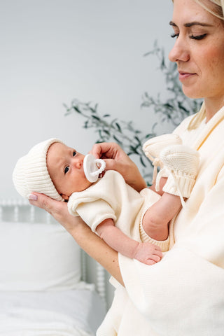 Lou Lou and Company Knit Booties - Ivory
Woman holding a baby wrapped in a white blanket with a white hat and pacifier.