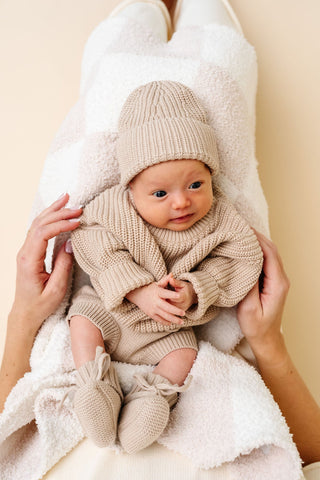 Lou Lou and Company Knit Booties - Tan
Baby wrapped in a beige knitted outfit with matching hat and booties, held by a person against a light background.