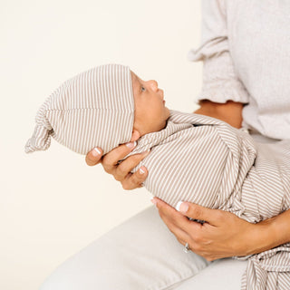 Lou Lou and Company Top Knot Hat - Banks
Newborn baby wrapped in a striped swaddle and cap being held by a person on a light background