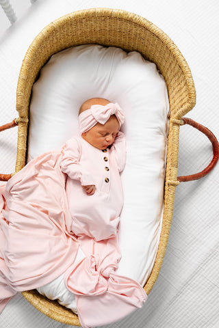 Lou Lou and Company Knotted Gown - Sophie
Newborn baby in a pink outfit and headband sleeping in a wicker crib with white bedding.