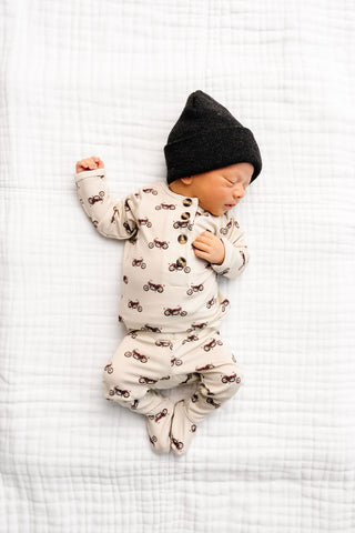 Lou Lou and Company Top and Bottoms - Cruz
Newborn baby sleeping on a white textured blanket wearing a black hat and patterned onesie.
