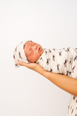 Lou Lou and Company Top Knot Hat - Cruz
Newborn baby swaddled in a patterned blanket held by a person against a white background