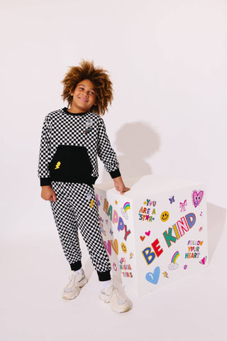 Lola and The Boys Checker Astro Patch Set
Person wearing a black and white checkered outfit with a colorful 'Be Kind' sign in the background.