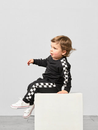 Little Bipsy Stripe Checkered Jogger - Black
Child wearing a black outfit with checkered details sitting on a white block against a light gray background