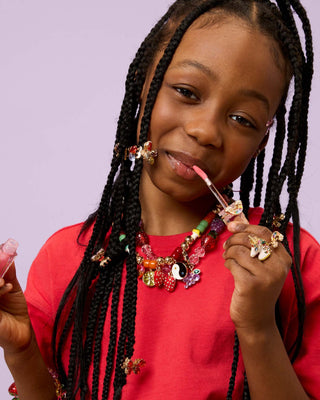 Super Smalls Gloss It Rainbow Lip Gloss Ring Set
Young girl with braided hair wearing colorful jewelry, holding a pink object.