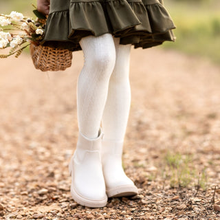 Little Stocking Co Cable Knit Tights - Ivory
Person wearing a green dress, white tights, and white shoes holding flowers on a gravel path.