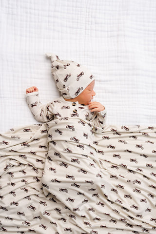 Lou Lou and Company Top Knot Hat - Cruz
Baby wrapped in a patterned blanket and wearing a matching hat on a white textured background