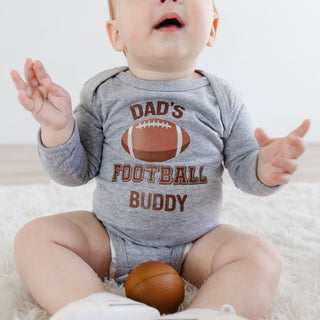 Sweet Wink Dad's Football Buddy Long Sleeve Bodysuit - Gray Baby wearing a gray onesie with 'Dad's Football Buddy' text and graphics, sitting on a white surface.