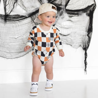 Sweet Wink Boo Checkered Halloween Long Sleeve Romper - Natural Baby wearing a checkered outfit with Halloween-themed design, standing against a stylized black and white background.