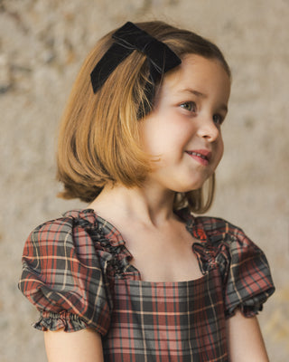 Noralee Louella Dress - Winter Tartan
Young girl wearing a plaid dress with a black bow in her hair against a textured wall.