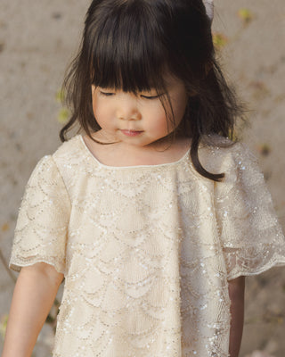 Noralee Daisy Dress - Silver Metallic
Young girl wearing a beige sequined dress against a blurred natural background