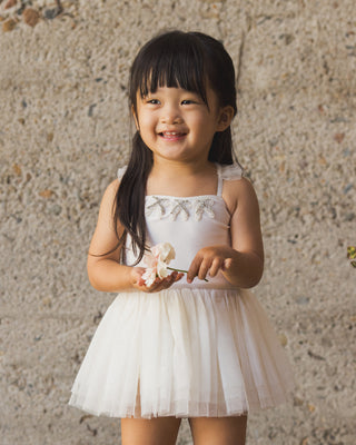 Noralee Clementine Tutu - Ivory
Young girl in a white dress holding a flower against a textured wall.