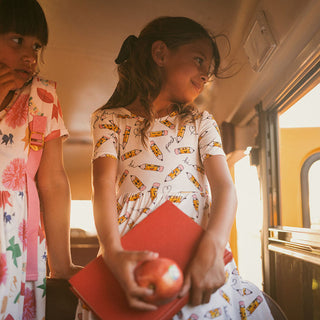 Posh Peanut Posh Pencils Ruffled Twirl Dress Two children on a school bus, one holding an apple and a red book.