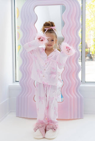 Lola and The Boys Kid's Pretty in Bows Silky Set Child in a pink and white outfit standing in front of a decorative mirror with wavy pink and purple design.