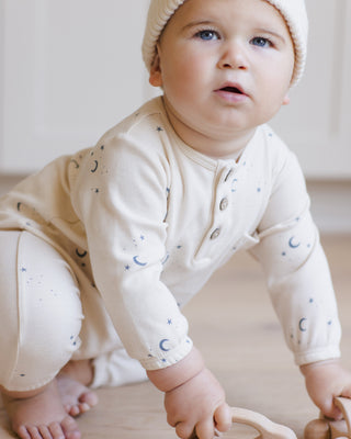 Quincy Mae Henley Bubble Jumpsuit - Moons
Baby wearing a cream outfit with star patterns and a matching hat, sitting on a light-colored floor.