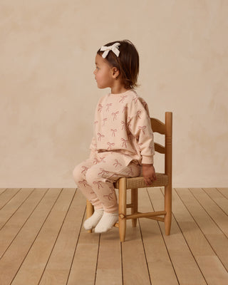 Quincy Mae Relaxed Fleece Sweatshirt & Sweatpant Set - Red Bows
Child wearing a pink outfit with bow pattern sitting on a wooden chair against a beige wall.