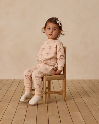 Quincy Mae Relaxed Fleece Sweatshirt & Sweatpant Set - Red Bows
Young child wearing a pink outfit with bow patterns, sitting on a wooden chair against a beige wall.