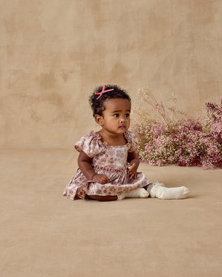 Noralee Margot Romper - Harvest Fleur
Baby girl in a floral dress sitting on a beige background with pink flowers.