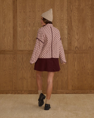 Rylee & Cru Womens Quilted Jacket - Blush Fleur Person wearing a patterned jacket and maroon skirt against a wooden paneled wall.