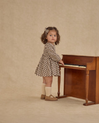 Rylee & Cru Capri Dress - Harvest Floral
Young girl in a patterned dress standing next to a wooden piano against a beige background