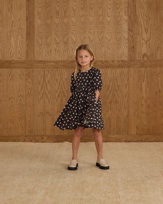 Rylee & Cru Rose Dress - Hearts
Young girl wearing a black dress with white patterns against a wooden paneled wall.