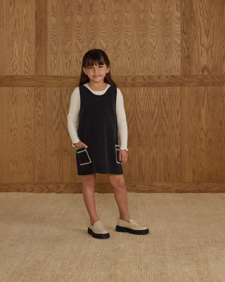 Rylee & Cru V-Neck Overall Dress - Washed Black
Young girl wearing a black dress with white sleeves against a wooden paneled wall.
