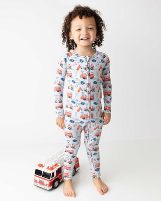 Lev Baby Poppy Convertible Zipper Romper - Ryan
Child wearing pajamas with vehicle pattern, standing next to a toy fire truck on a white background