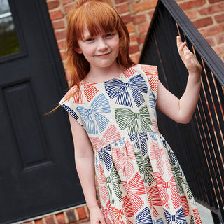 Pink Chicken Girls Adaline Dress - Multi Striped Bows Young girl wearing a colorful dress with bow pattern standing in front of a door.