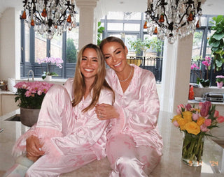 Lola and The Boys Women's Pretty in Bows Silky Set Two women in pink satin pajamas sitting together in a stylish room with chandeliers and flowers.