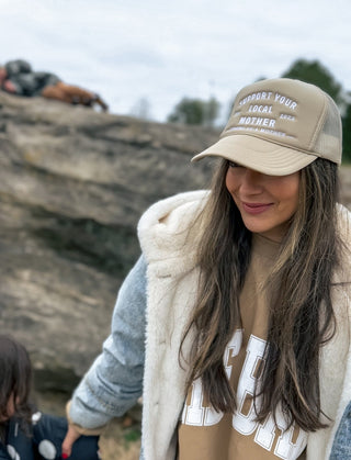 Support Your Local Mother® Trucker Hat - Tan
Woman wearing a cap with text, standing outdoors near rocks