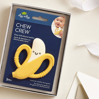 Itzy Ritzy Chew Crew® - Banana Yellow silicone teether with banana design in a 'Itzy Ritzy' packaging on a beige surface.