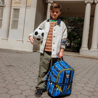 State Bags Kane Double Pocket Backpack - Soccer Jersey Child holding a soccer ball and a blue backpack in front of a building
