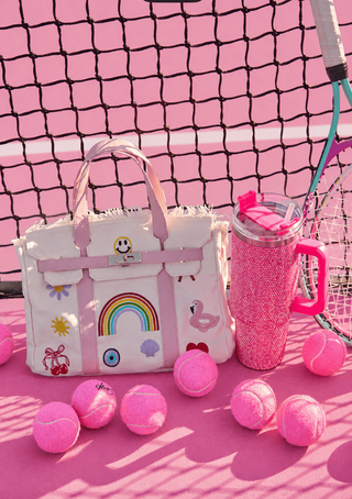 Lola and The Boys Beachkin Bag
Pink tennis bag with rainbow and cloud design, next to pink tennis balls and a pink tumbler on a pink tennis court.